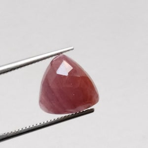 Puede incluir: Una gema triangular facetada, de color rojo intenso. La gema está sujeta por unas pinzas plateadas, mostrando sus superficies pulidas y su claridad interna. La forma de la piedra es un triángulo modificado.