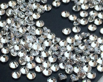 May include: A close-up view of a large quantity of round, clear, faceted gemstones. The gemstones are scattered on a black surface.