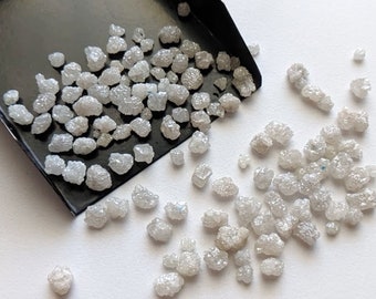 May include: A close-up of a pile of small, rough, gray diamonds. The diamonds are scattered on a white surface and a black scoop.