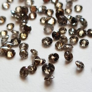 Può includere: Una collezione di piccoli diamanti neri rotondi sparsi su una superficie bianca. I diamanti hanno una varietà di dimensioni e forme, e sono tutti molto scintillanti.