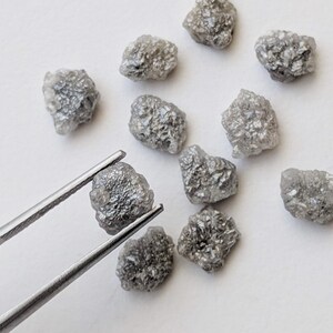 K&ouml;nnte beinhalten: Eine Sammlung von zehn rohen, grauen Diamanten, von denen einige mit einer Pinzette gehalten werden. Die Diamanten sind in verschiedenen Gr&ouml;&szlig;en und Formen.