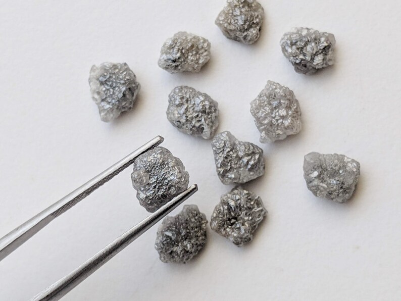 K&ouml;nnte beinhalten: Eine Sammlung von zehn rohen, grauen Diamanten, von denen einige mit einer Pinzette gehalten werden. Die Diamanten sind in verschiedenen Gr&ouml;&szlig;en und Formen.