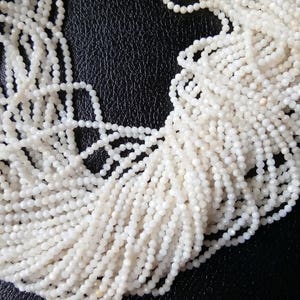 May include: A strand of white gemstone beads, each bead is faceted and has a slightly iridescent sheen. The beads are strung together in a long strand, and the strand is coiled up on a dark surface.