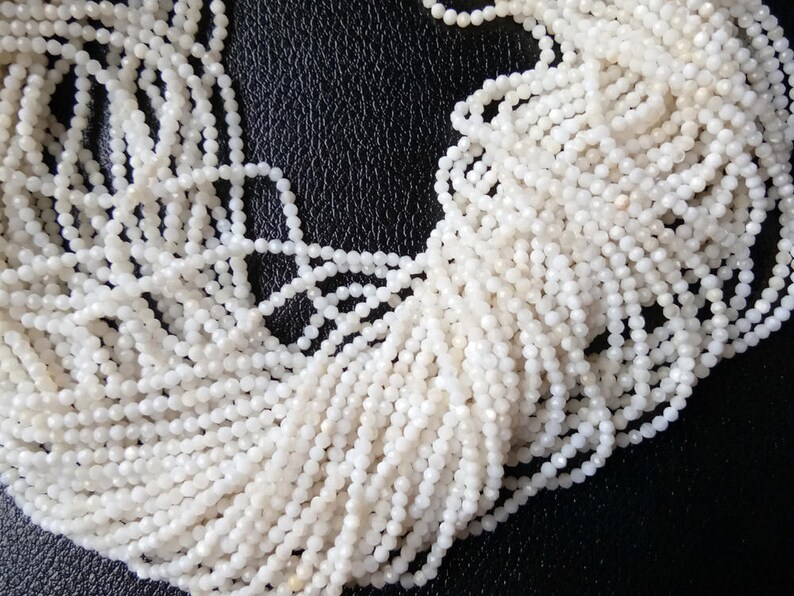 May include: A strand of white gemstone beads, each bead is faceted and has a slightly iridescent sheen. The beads are strung together in a long strand, and the strand is coiled up on a dark surface.