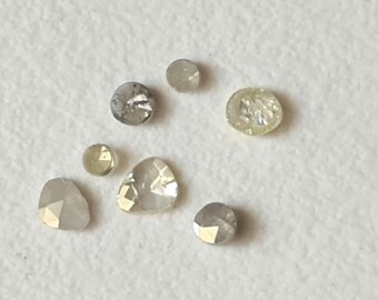 May include: An assortment of small, uncut diamonds in various shapes and colors, including round, pear, and irregular forms. The diamonds range in color from clear to gray and yellow, with visible facets.