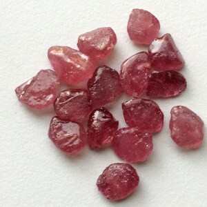 May include: A collection of 15 rough, pink ruby gemstones. The stones are various sizes and shapes, with some having a more rounded shape and others being more angular.