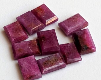May include: A collection of nine rectangular, faceted rubies with a deep magenta and crimson hue. Each gemstone displays a polished surface with visible natural inclusions and internal textures, arranged in a loose cluster on a white surface.