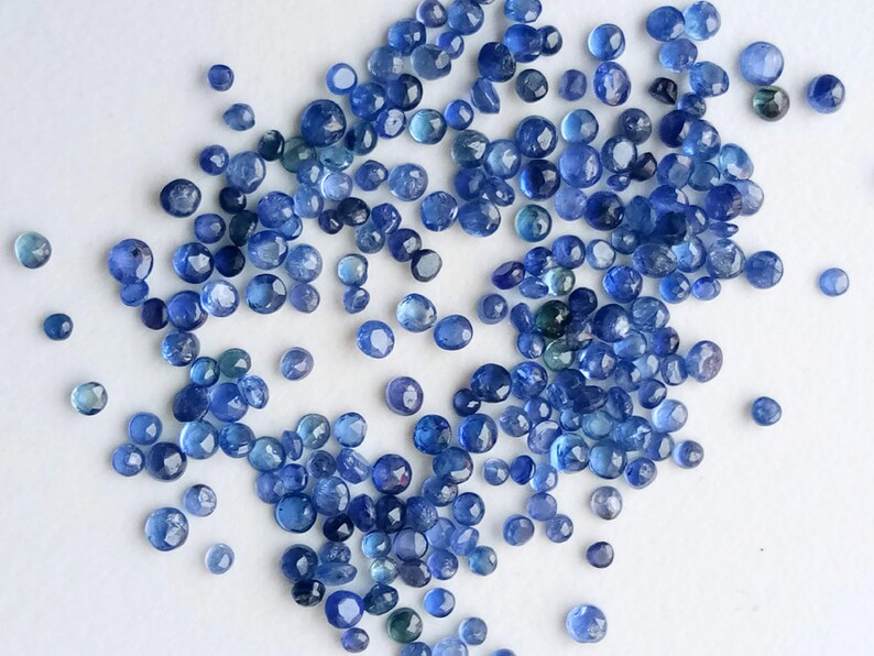 May include: A large group of blue gemstone beads, scattered on a white surface. The beads are round and have a slightly irregular shape.