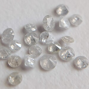 May include: A collection of small, round, white diamonds with a rough, unpolished finish. The diamonds are scattered on a white surface.