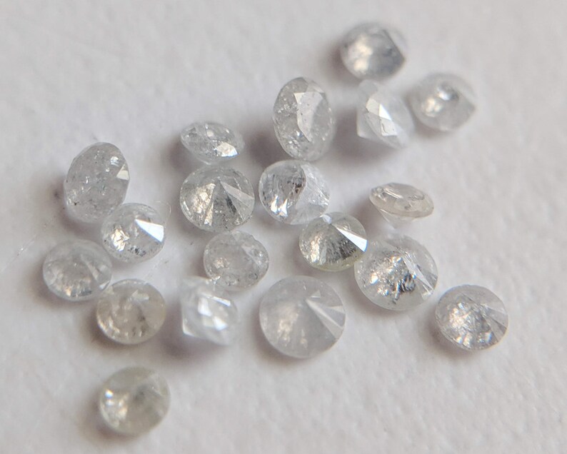 May include: A collection of small, round, white diamonds with a rough, unpolished finish. The diamonds are scattered on a white surface.