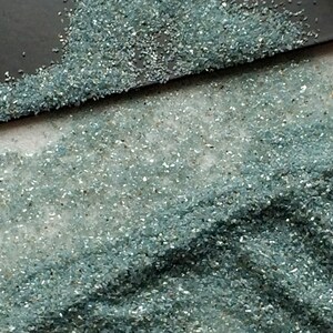 May include: A close-up of a pile of small, light blue glass pieces. The glass pieces are scattered on a white surface.