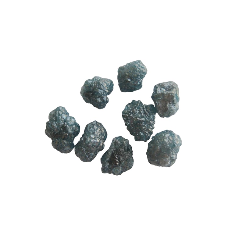 May include: A group of nine dark gray, rough, and unpolished diamonds. The diamonds are scattered on a white background.