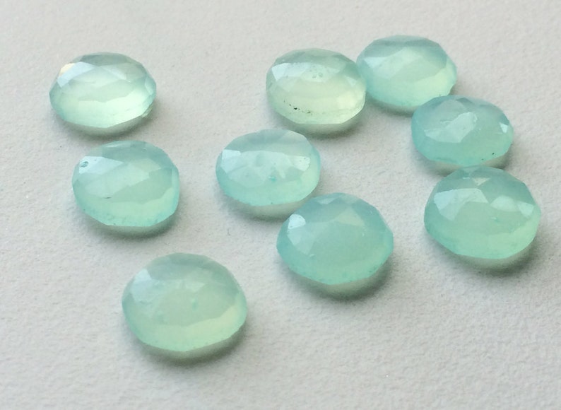 May include: A collection of ten round, faceted gemstones in shades of pale blue-green. The gemstones have a polished, translucent appearance and are arranged on a light-colored surface. Each stone is approximately 0.5 inches in diameter.