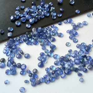 May include: A loose pile of small, round, blue gemstones. The gemstones are scattered on a white surface.