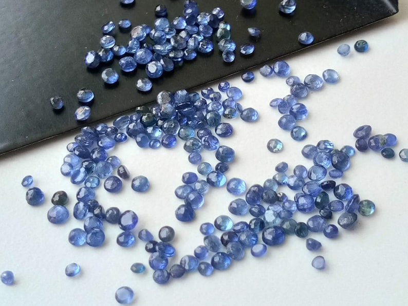 May include: A loose pile of small, round, blue gemstones. The gemstones are scattered on a white surface.