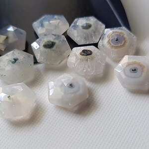 May include: Ten white, faceted, hexagonal gemstone beads with dark gray and brown inclusions. The beads are arranged in a cluster on a white surface.