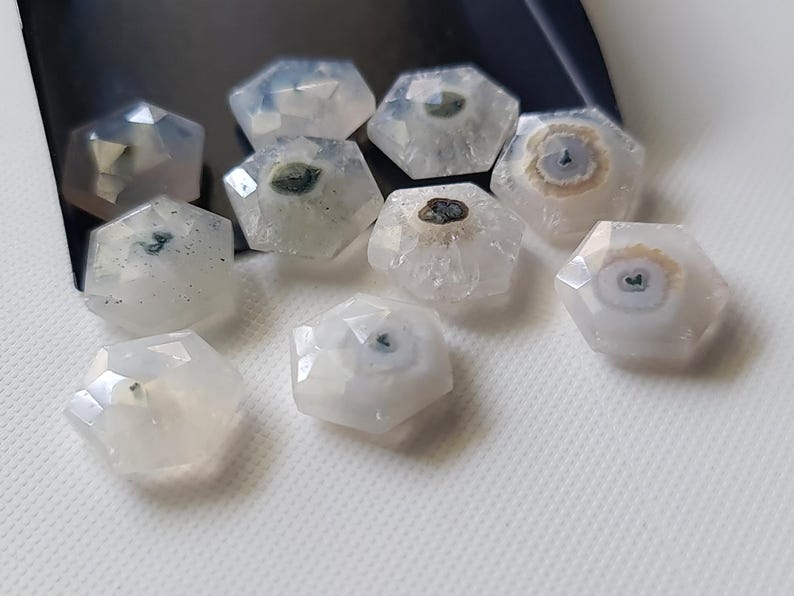 May include: Ten white, faceted, hexagonal gemstone beads with dark gray and brown inclusions. The beads are arranged in a cluster on a white surface.