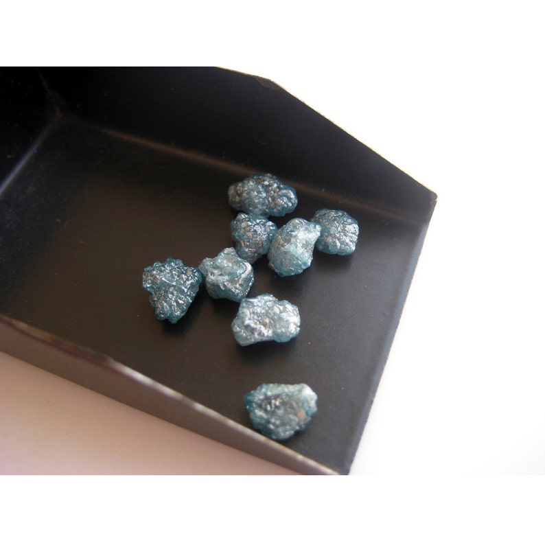 May include: A small black tray holds a collection of rough, blue-grey diamonds. The diamonds are various sizes and shapes, with some having a more crystalline appearance.