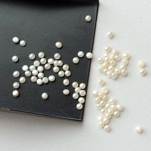 May include: A close-up of a collection of white round pearls, some on a black tray and others scattered on a white surface.