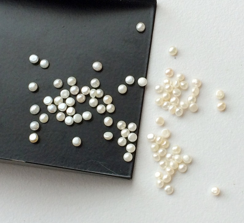 May include: A close-up of a collection of white round pearls, some on a black tray and others scattered on a white surface.