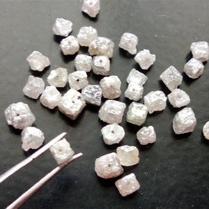 May include: A collection of rough, white, diamond crystals, some of which are faceted, are scattered on a dark gray surface. A pair of tweezers holds a single, small, white diamond crystal.