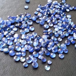 May include: A large quantity of small, faceted, blue gemstones. The gemstones are scattered on a dark grey surface.