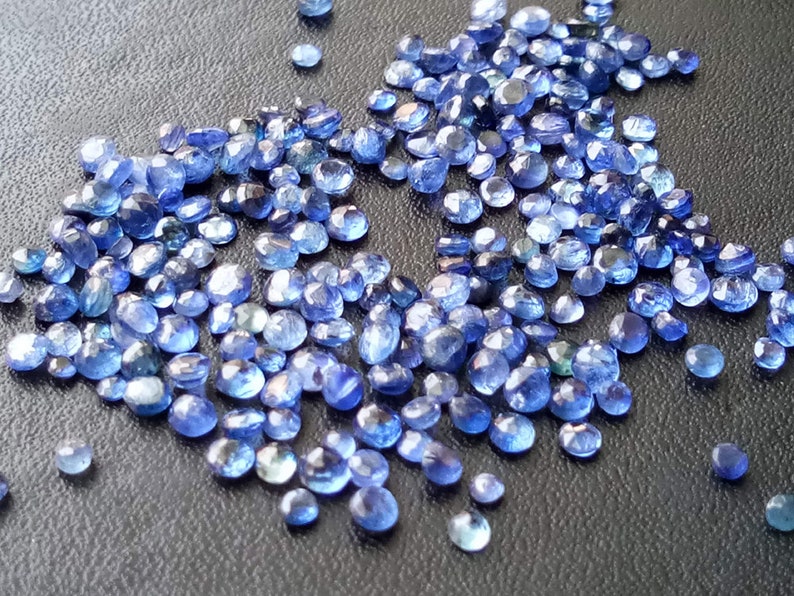 May include: A large quantity of small, faceted, blue gemstones. The gemstones are scattered on a dark grey surface.