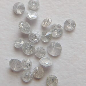 May include: A collection of small, round, clear, faceted diamonds. The diamonds are scattered on a white surface.