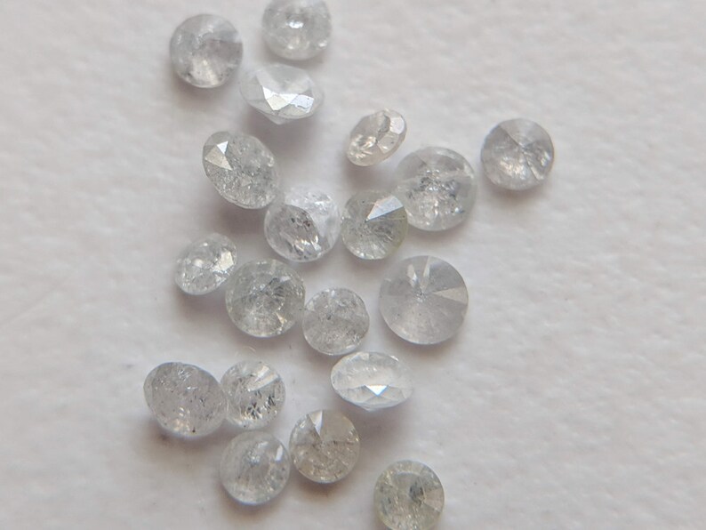 May include: A collection of small, round, clear, faceted diamonds. The diamonds are scattered on a white surface.