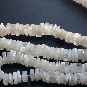 Puede incluir: Una hebra de cuentas de piedra lunar blanca, cada cuenta es un prisma rectangular con una forma ligeramente irregular. Las cuentas tienen un brillo iridiscente sutil.