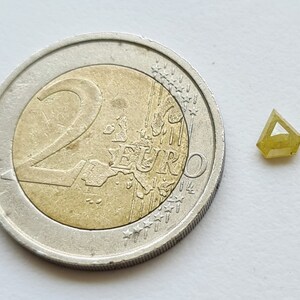 May include: A small, triangular, yellow diamond gemstone next to a 2 Euro coin for size comparison.