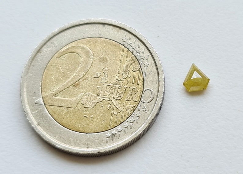 May include: A small, triangular, yellow diamond gemstone next to a 2 Euro coin for size comparison.