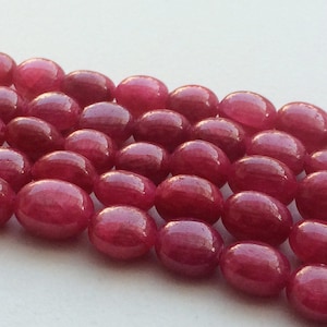 Puede incluir: Una hebra de cuentas de piedras preciosas ovaladas de color rosa intenso. Las cuentas son lisas y brillantes, con un ligero brillo.