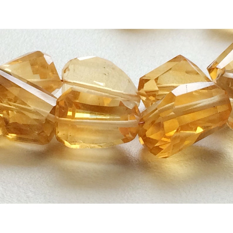 May include: A close-up of a strand of faceted, golden citrine beads. The beads are cut into a variety of shapes, including round, oval, and square. The beads are strung on a clear thread.