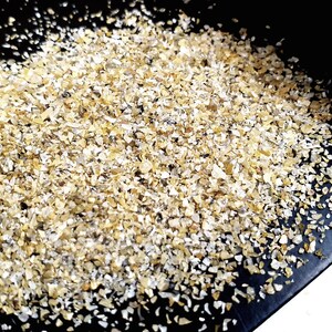 May include: A close-up of a pile of small, light brown and white flakes of oat groats on a black surface.