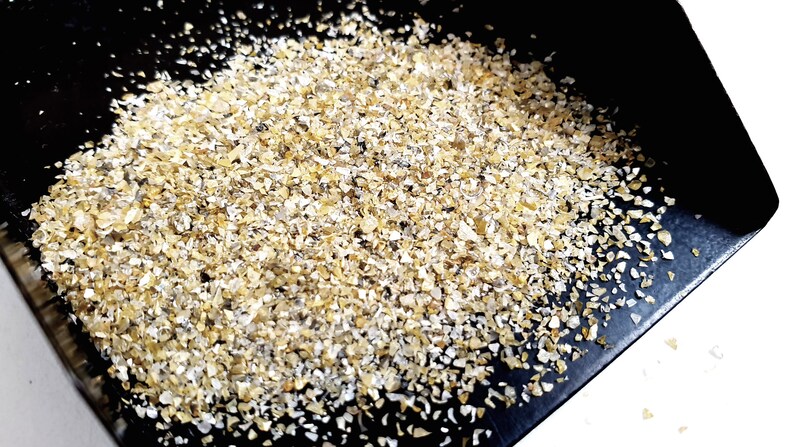 May include: A close-up of a pile of small, light brown and white flakes of oat groats on a black surface.
