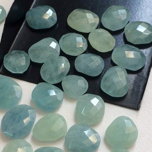 May include: A collection of light blue aquamarine gemstones, faceted and polished, in various sizes and shapes. The stones are arranged on a black surface and a white surface.