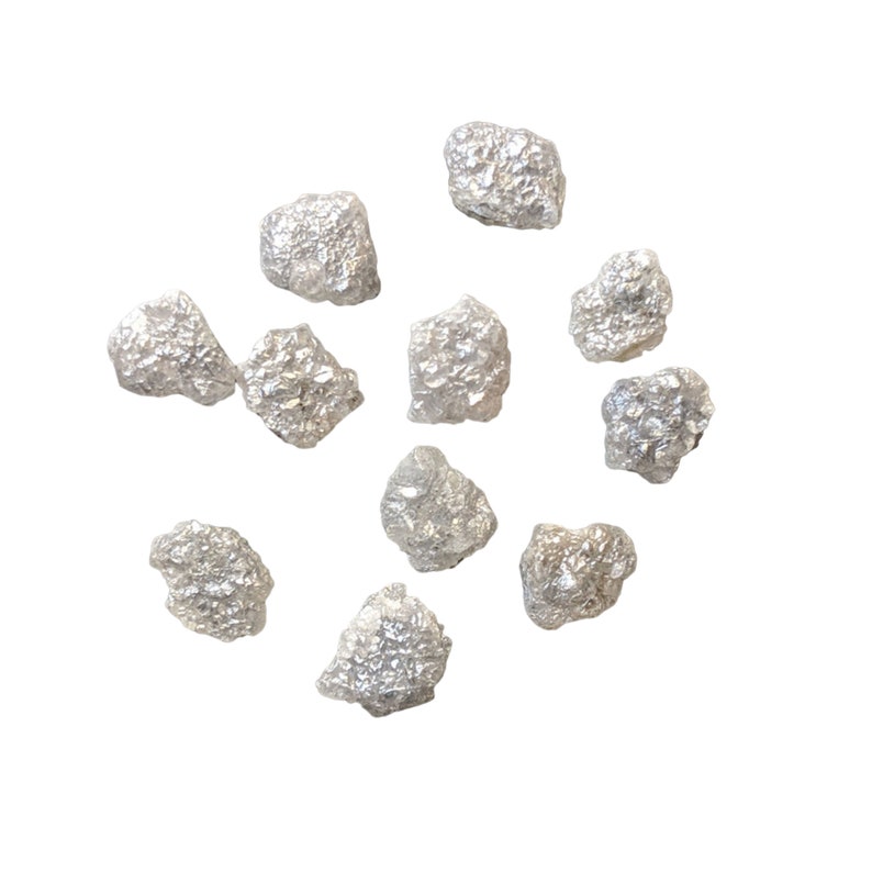 K&ouml;nnte beinhalten: Eine Sammlung von rohen, unpolierten, grauen Diamanten. Die Diamanten sind auf einem wei&szlig;en Hintergrund verstreut.