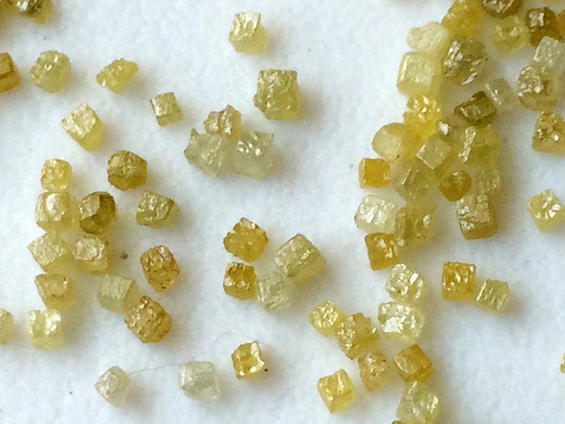 May include: A collection of small, yellow-green, faceted, cubic crystals. The crystals are scattered on a white surface.