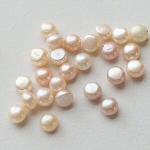 May include: A collection of small, round, white and pink pearls. The pearls are scattered on a white surface.