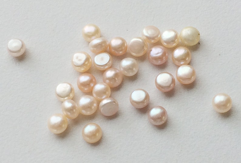 May include: A collection of small, round, white and pink pearls. The pearls are scattered on a white surface.