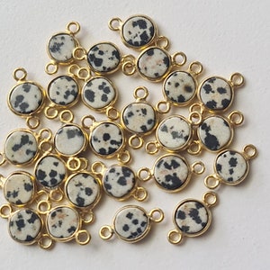 Pode incluir: Uma coleção de 20 pingentes de metal dourado com detalhes de pedra de jaspe dalmata redonda. Cada pingente tem dois anéis de salto para prender em joias.