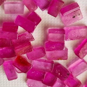 Puede incluir: Una colección de piedras preciosas rectangulares facetadas de color rosa. Las piedras preciosas están dispuestas en un patrón aleatorio sobre una superficie blanca.