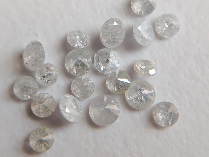 May include: A collection of small, round, white diamonds with a rough, uncut finish. The diamonds are scattered on a white surface.