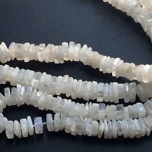 Puede incluir: Una hebra de cuentas de piedra lunar blanca, cada cuenta tiene una forma rectangular con un borde ligeramente irregular. Las cuentas est&aacute;n enhebradas para formar un collar.
