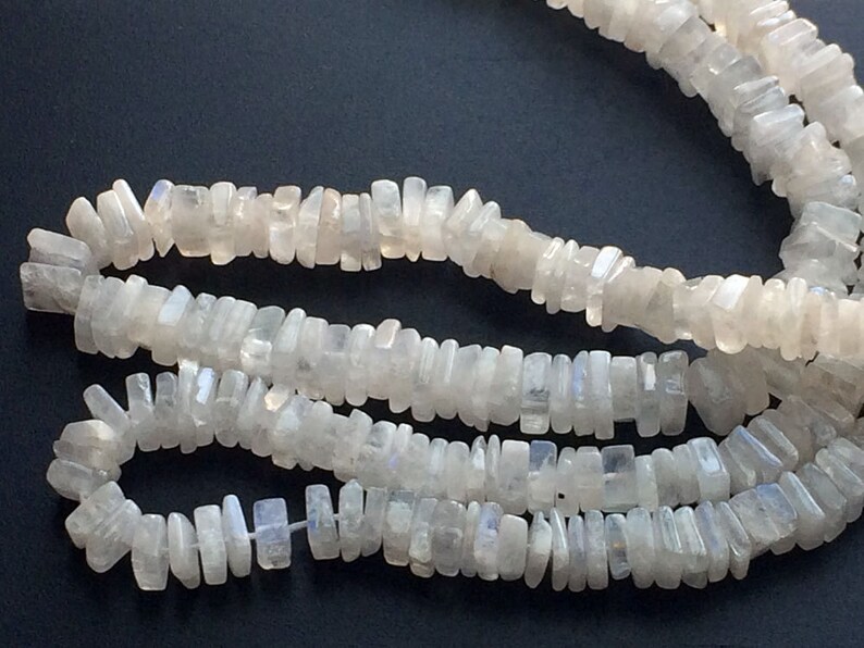 Puede incluir: Una hebra de cuentas de piedra lunar blanca, cada cuenta tiene una forma rectangular con un borde ligeramente irregular. Las cuentas est&aacute;n enhebradas para formar un collar.