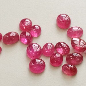 Pu&ograve; includere: Una collezione di 15 cabochon di rubino rosa, alcuni ovali e alcuni rotondi, su uno sfondo bianco.