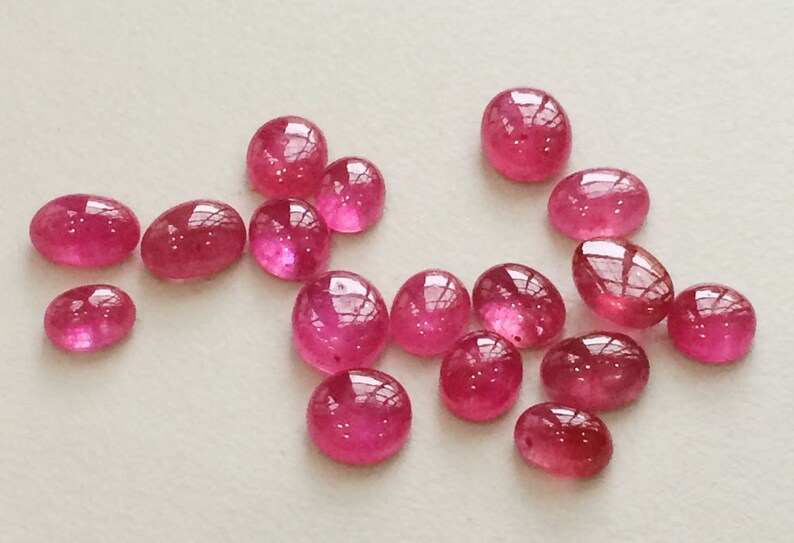 Pu&ograve; includere: Una collezione di 15 cabochon di rubino rosa, alcuni ovali e alcuni rotondi, su uno sfondo bianco.