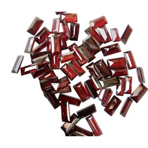 May include: A collection of loose, faceted, red gemstone rectangular beads. The beads are all approximately the same size and shape, and they are arranged in a random pattern.