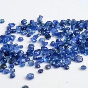 May include: A loose pile of small, faceted, blue gemstones. The gemstones are scattered on a white surface.